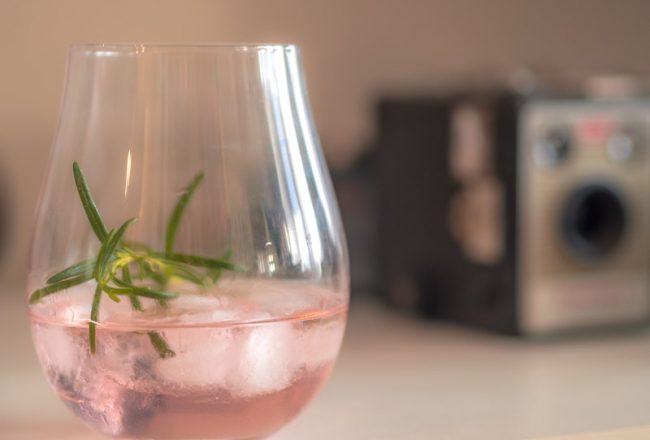 A glass is quarter filled with pin gin, ice-cubes and a sprig of rosemary. In the background is a vintage camera.