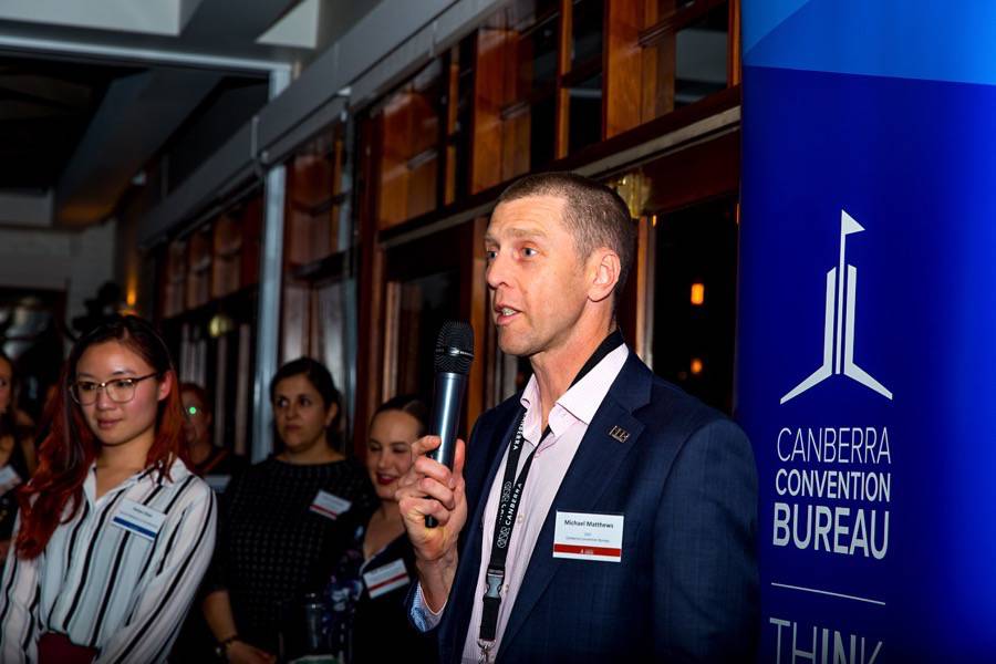 Man wearing suit holds a microphone and talks to a group of people. The sign behind him reads Canberra Convention Bureau.